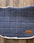 Sottosella da dressage trapuntato grigio-blu con profilo color crema, disteso su legno
