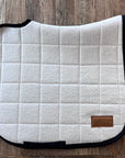 Sottosella da dressage trapuntato bianco naturale con profilo scuro, disteso su legno
