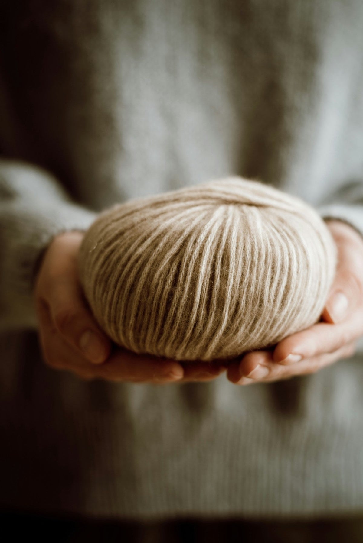 Hände halten ein Knäuel naturbeiger Wolle vor einem Strickpullover